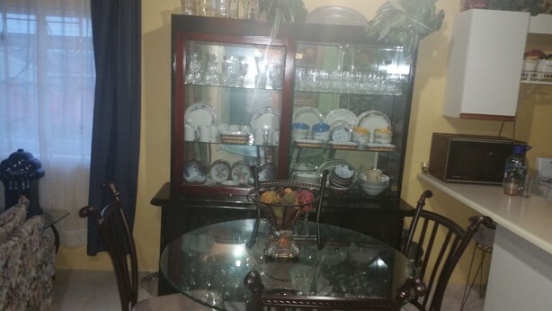 A dining area with a glass-top table surrounded by chairs, a glass cabinet with dishes, and a small television on a shelf. - Palm Cove - 2 Bedroom Cottage (Montego Bay)
