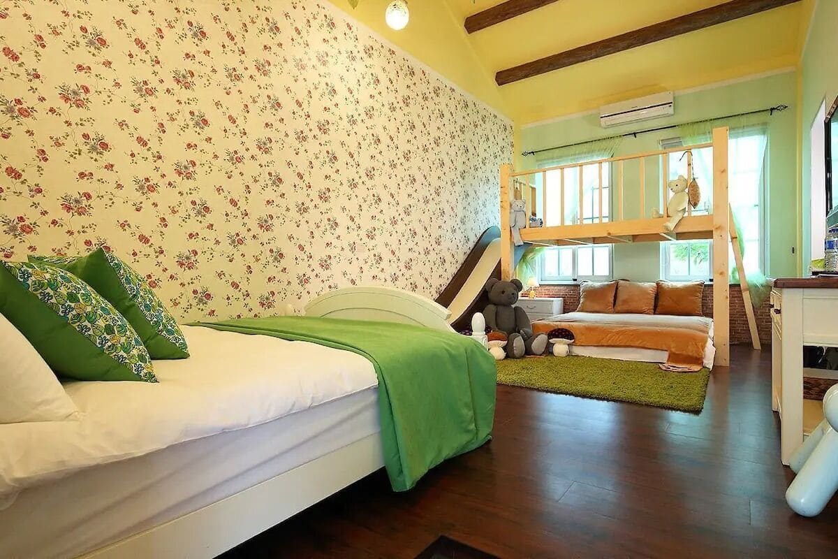 A cozy room with a bunk bed, a single bed with green pillows, floral wallpaper, wooden beams, and a window with a view.