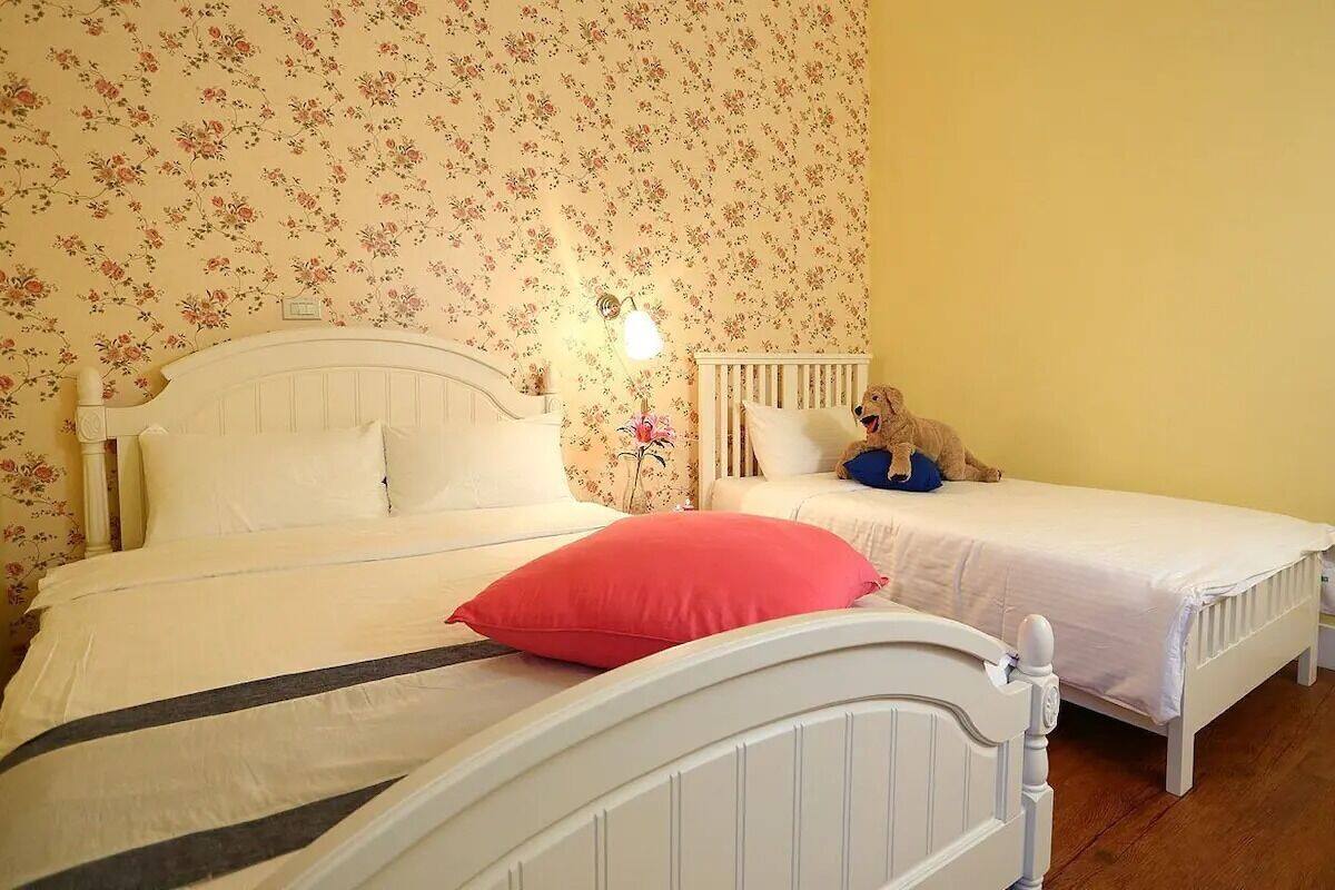 A room with two single beds, floral wallpaper, a nightstand with a lamp, and a teddy bear on one bed.
