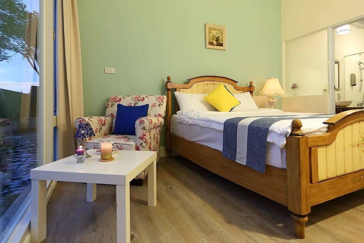 A bedroom with a wooden bed, a floral armchair, a bedside table with a candle, and a window with curtains.