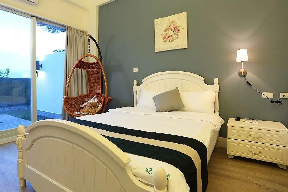 A bedroom with a bed, a nightstand, a wall-mounted lamp, a hanging chair, and a view of the outdoors.