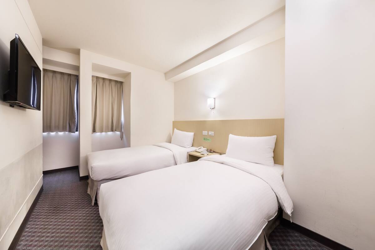 Habitación de hotel con dos camas, un televisor, una mesita de noche con teléfono y una ventana con cortinas.