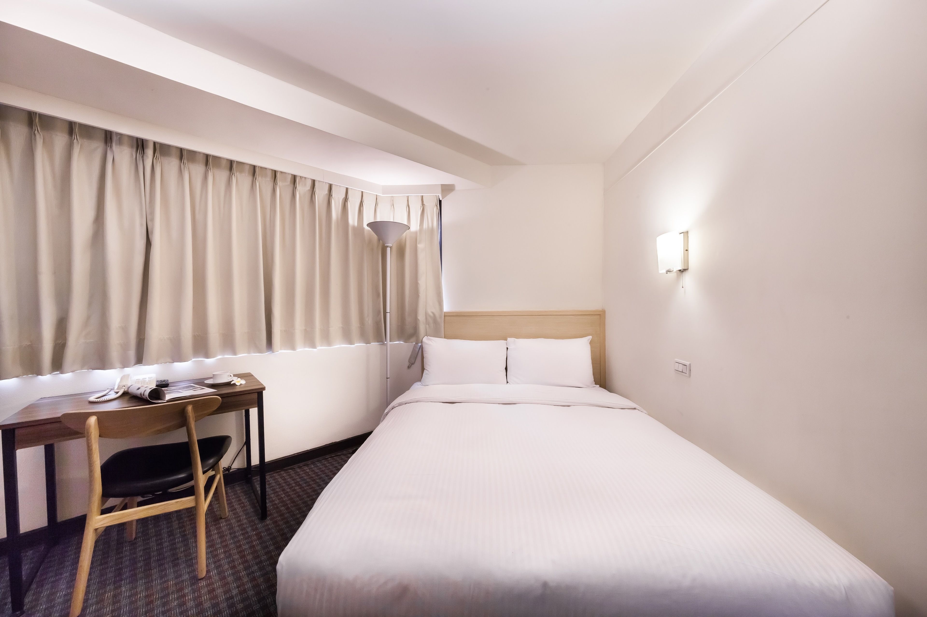 Habitació d'hotel amb llit, escriptori i cadira, una làmpada i una finestra amb cortines.
