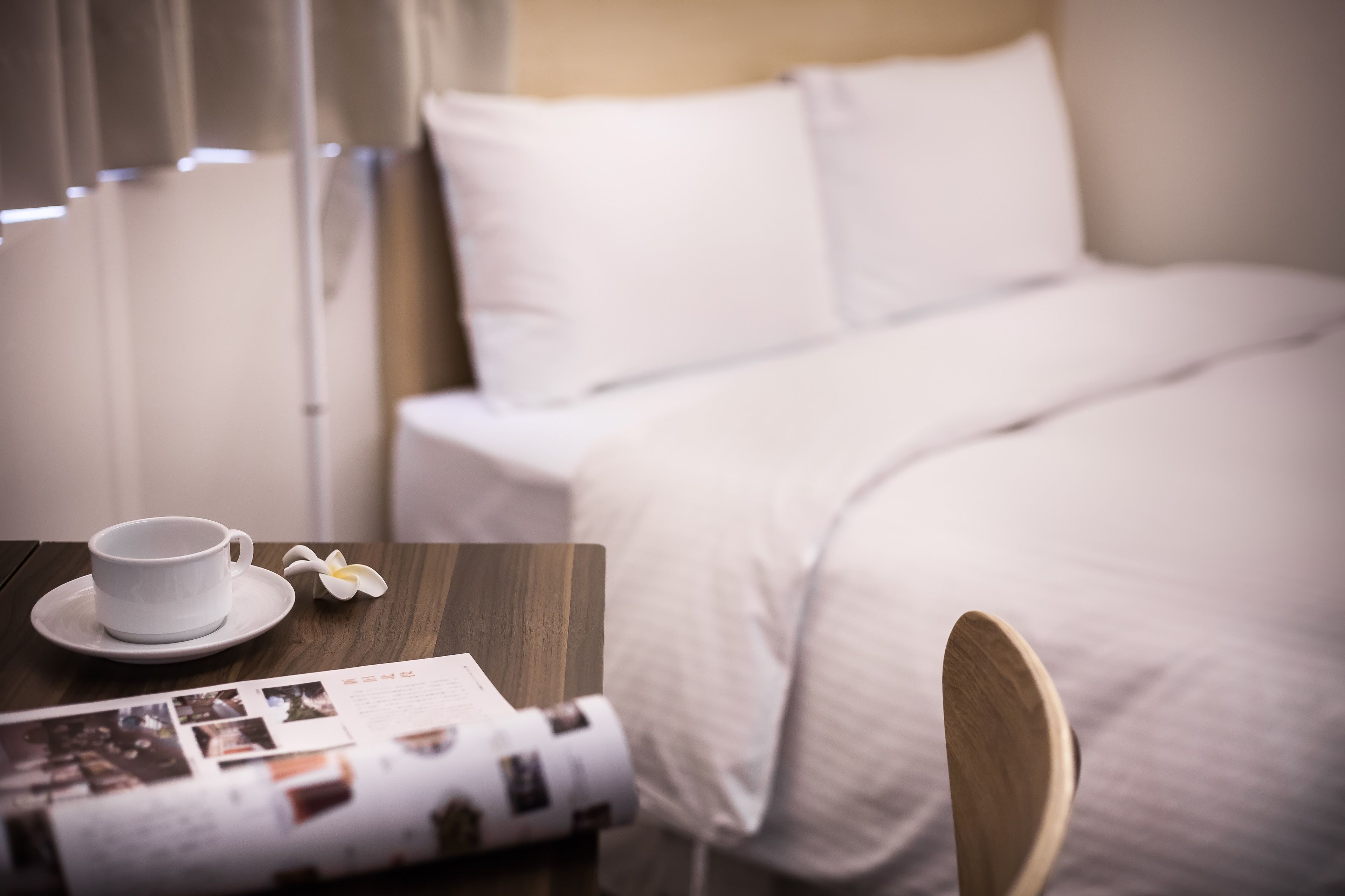 A neatly made bed with white bedding, a wooden nightstand with a cup and saucer, and a magazine.