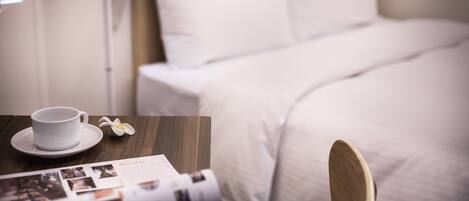 A neatly made bed with white bedding, a wooden nightstand with a cup and saucer, and a magazine.