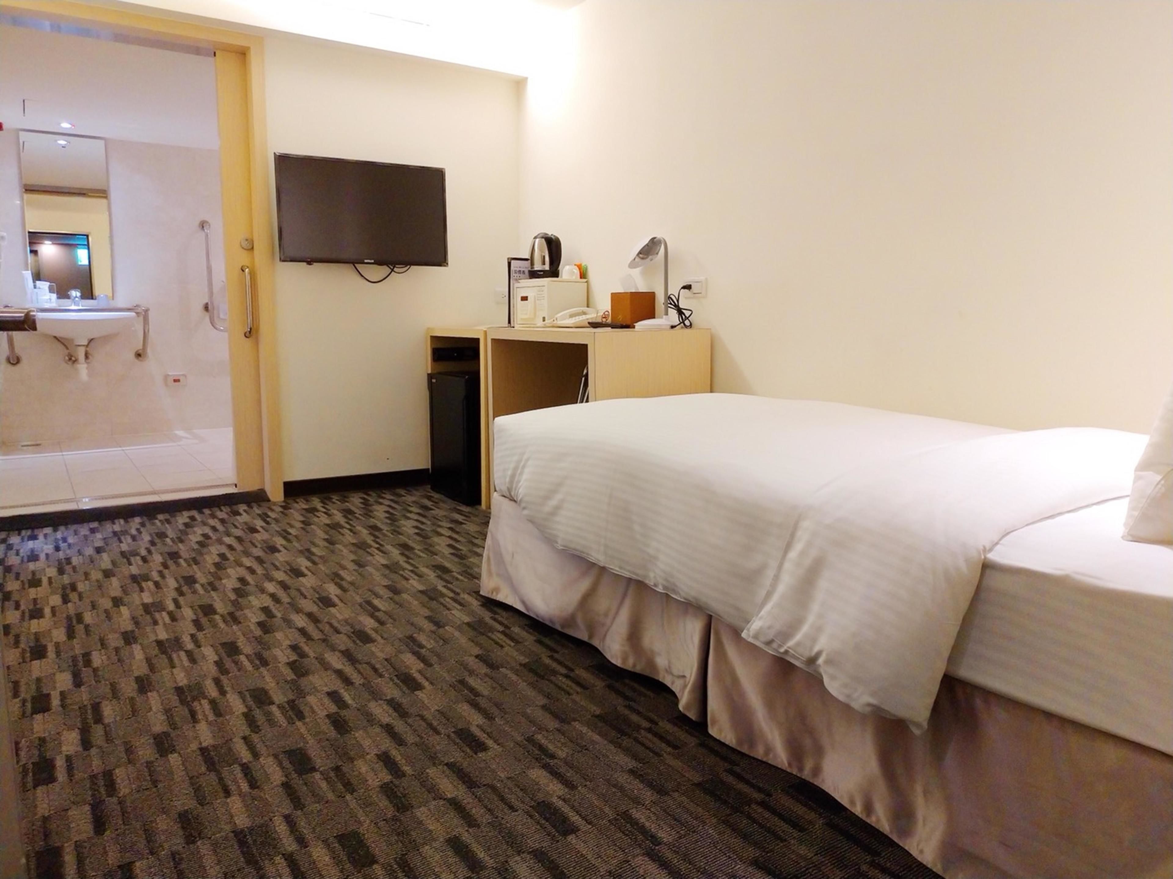 Ein Hotelzimmer mit Bett, Schreibtisch mit Lampe, Fernseher und Badezimmer mit Waschbecken und Spiegel.
