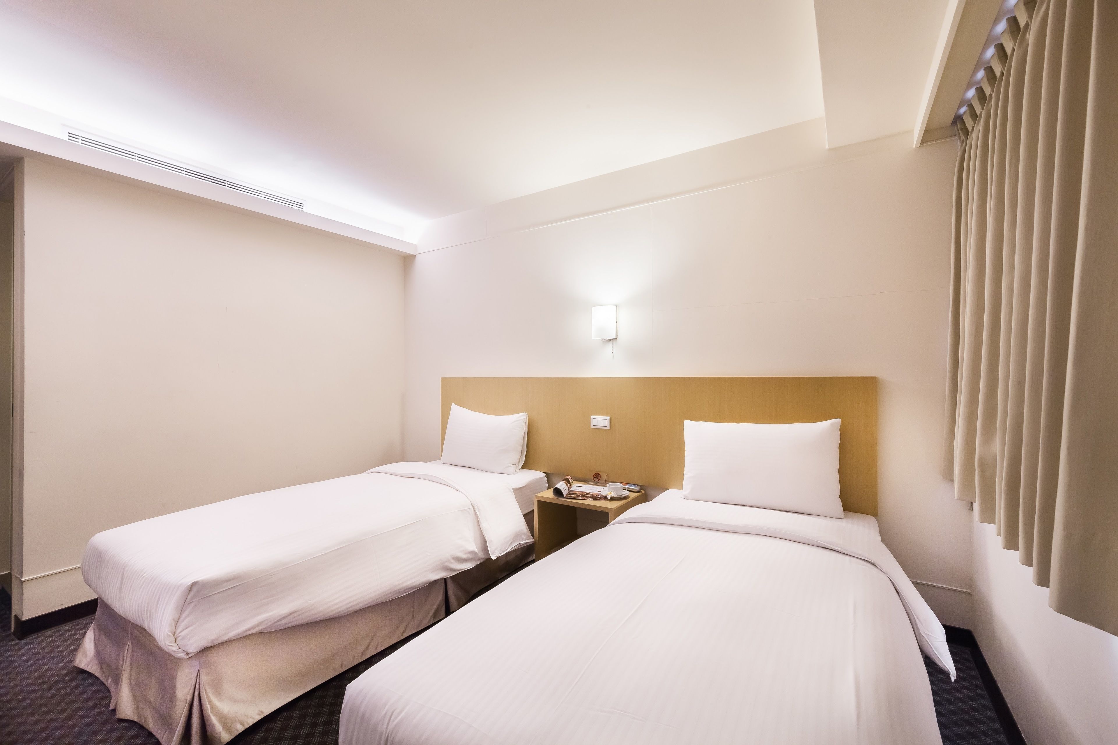 Une chambre d’hôtel avec deux lits simples, une tête de lit en bois, une table de chevet avec un téléphone et une lampe, et une fenêtre avec des rideaux.