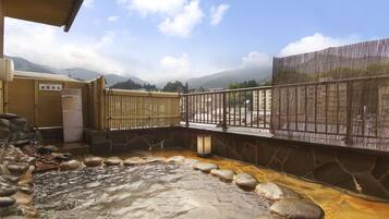 Un onsen japonés tradicional con un baño de piedra, escalones de madera y vistas a montañas.