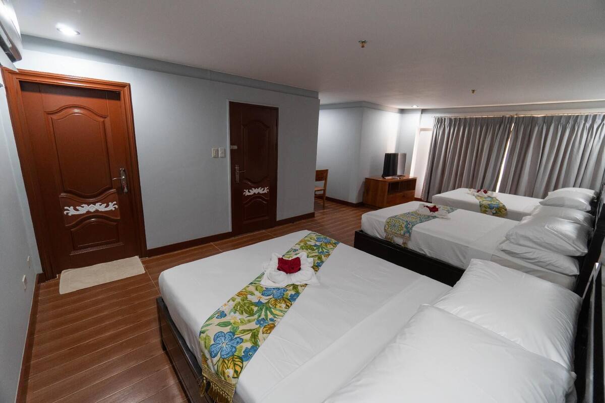 A hotel room with two beds, a wooden floor, a door, a small table with a lamp, and a curtain.