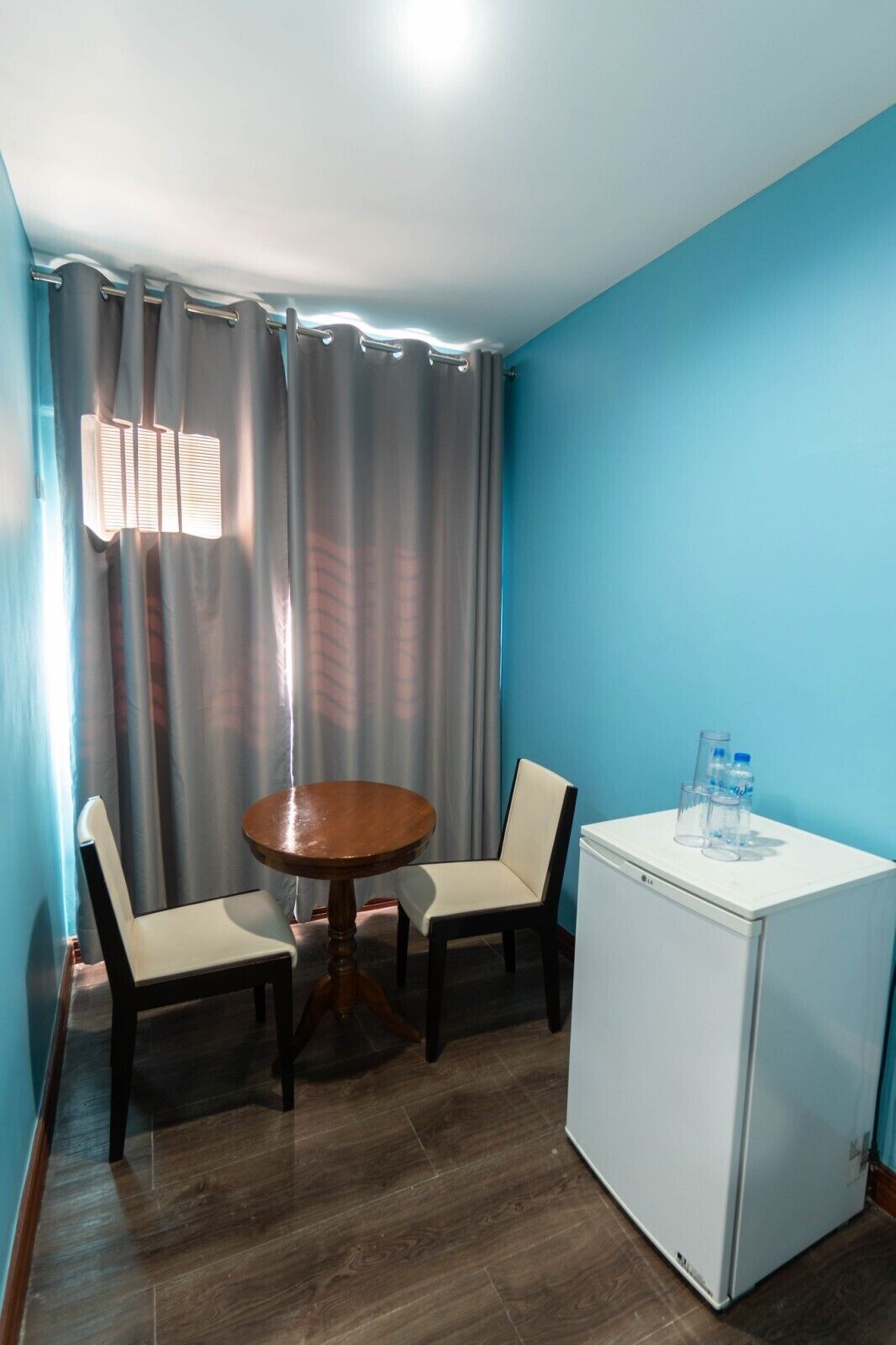A small room with a round wooden table, two chairs, a white refrigerator, and a window with curtains.