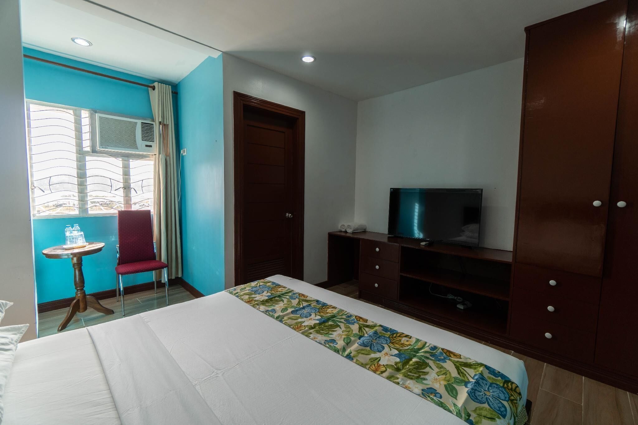 Een hotelkamer met een bed, een televisie, een kledingkast, een klein tafeltje met een stoel en een airconditioning.