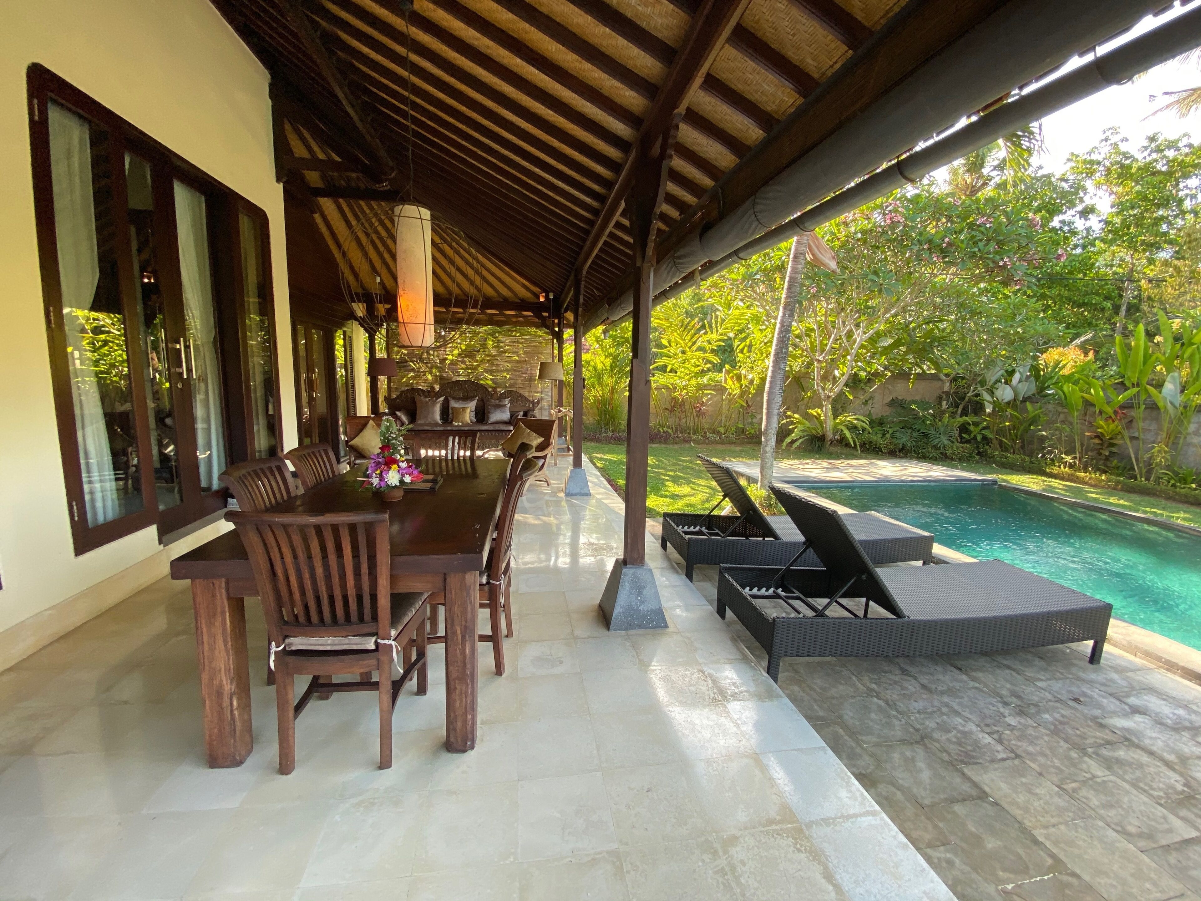 A covered outdoor area with a pool, wooden furniture, and a view of the surrounding greenery.