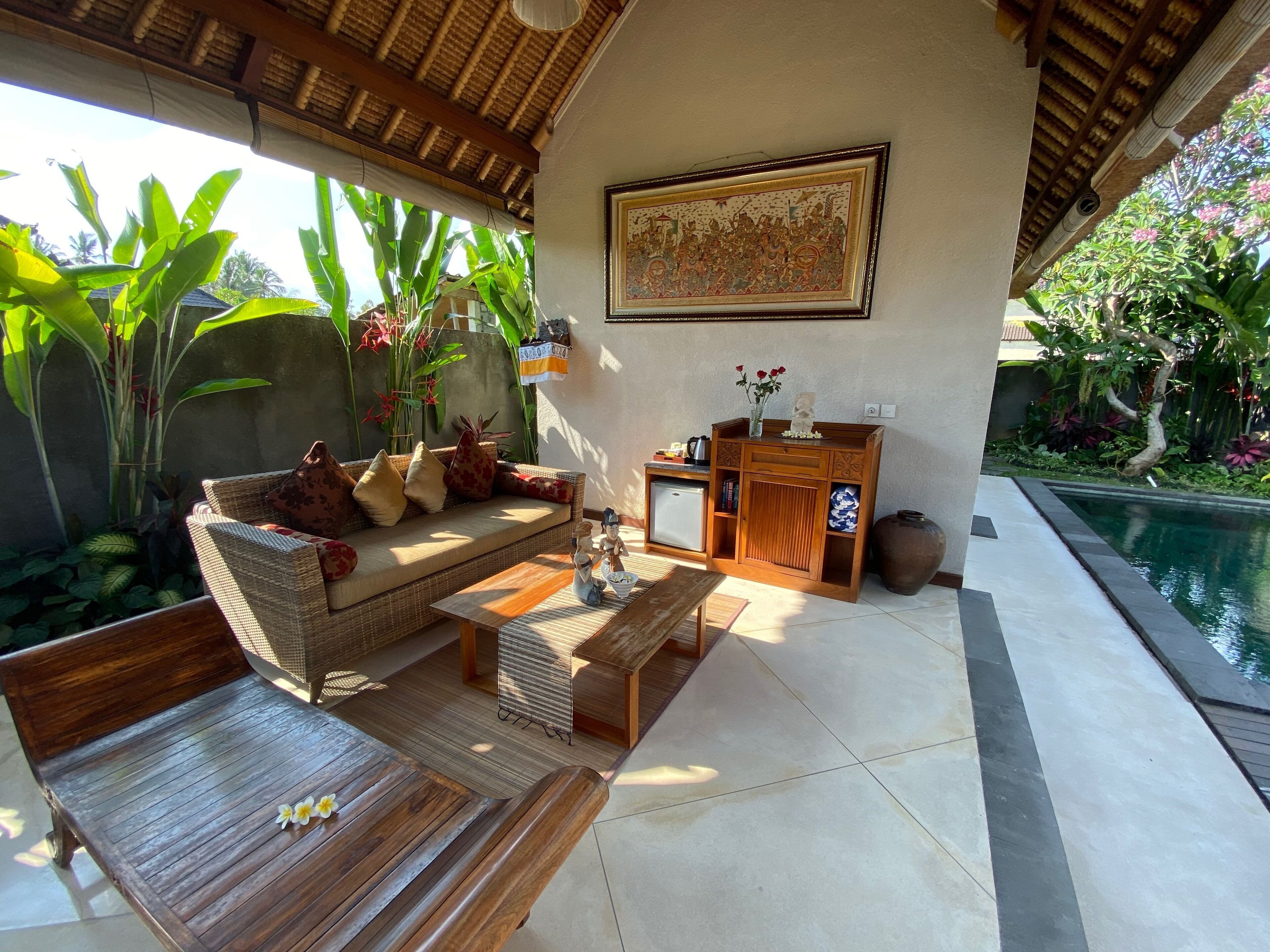A cozy outdoor living area with a sofa, coffee table, and dining table, surrounded by lush plants and a pool.