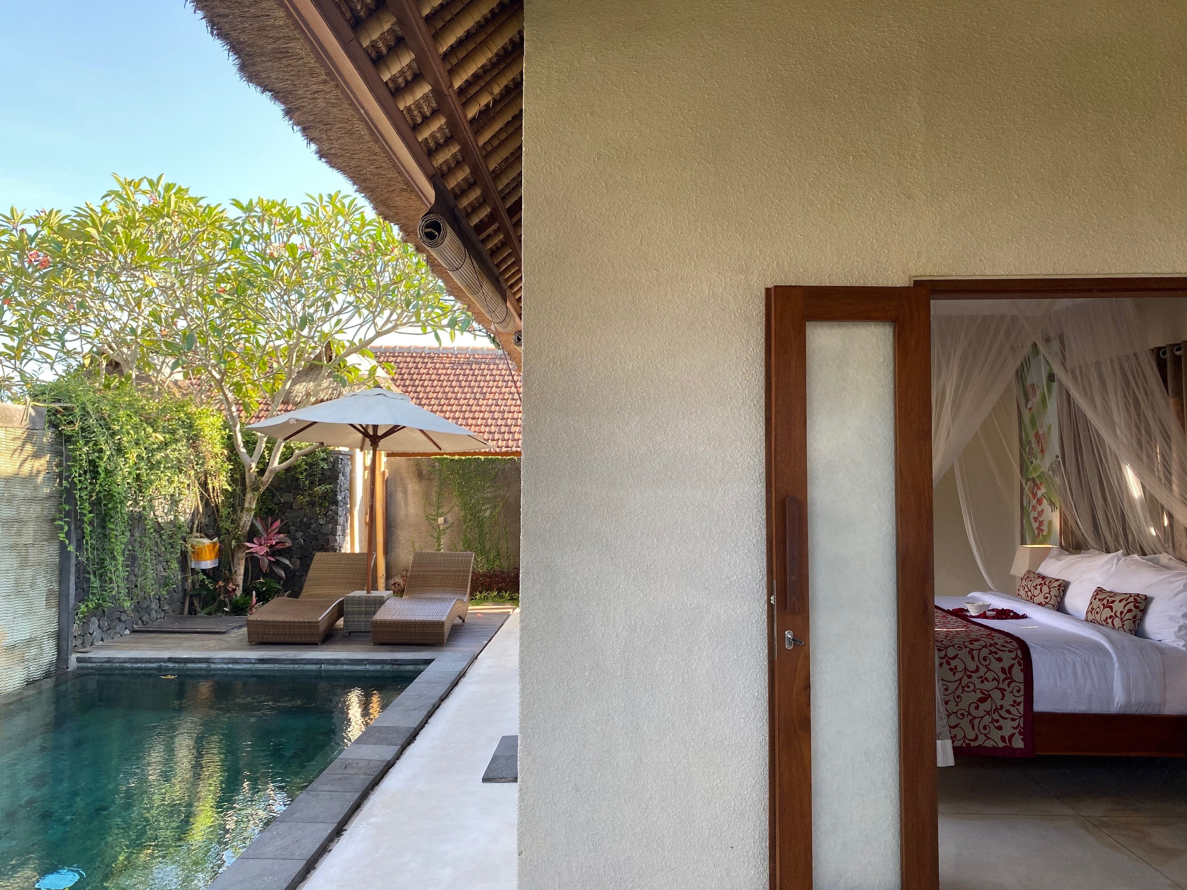 A bedroom with a canopy bed, a pool with lounge chairs, and a patio area with a table and umbrella.