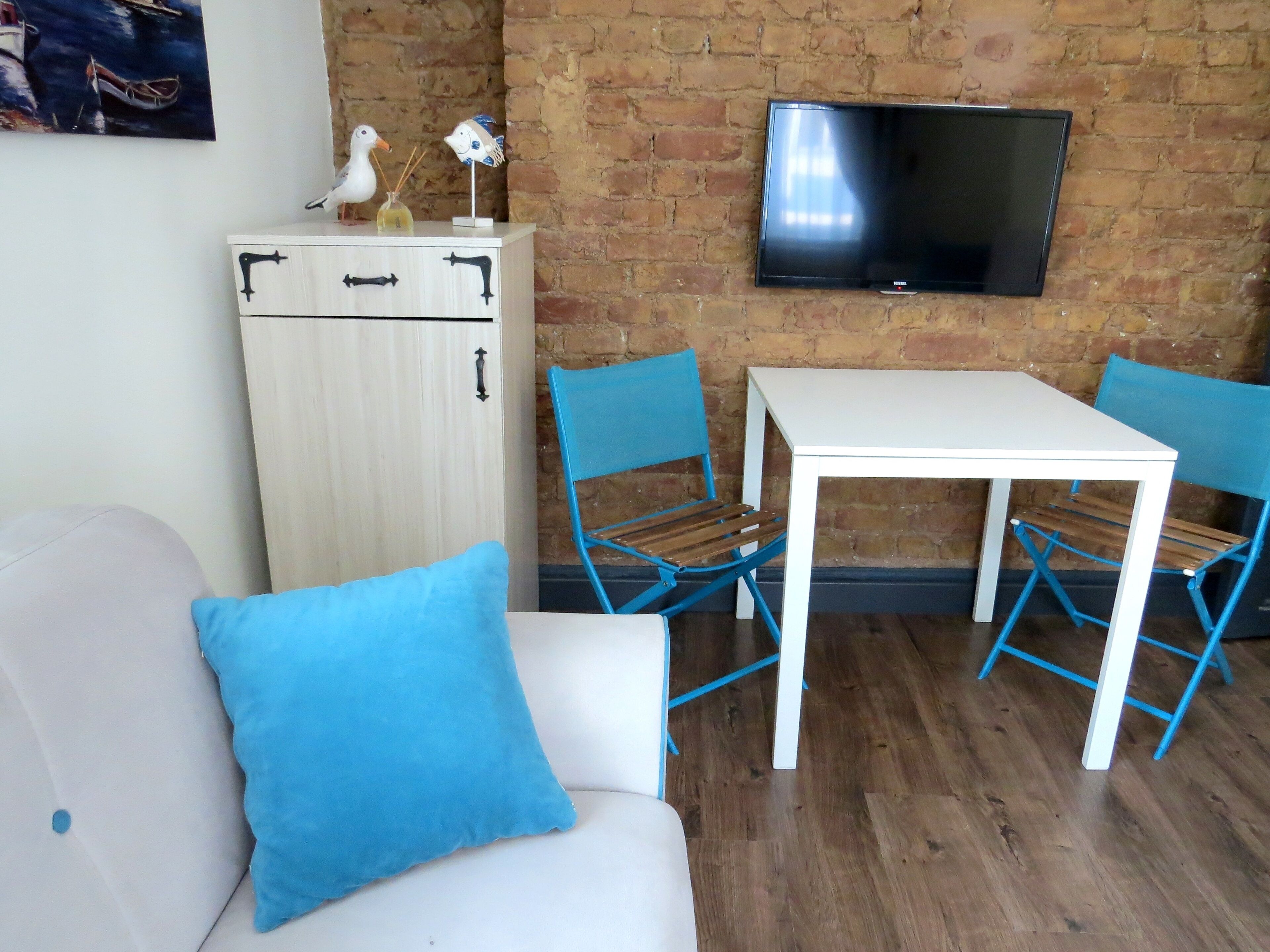 A compact living space with a white sofa, a blue cushion, a small table, two blue chairs, a white cabinet, and a flat-screen TV mounted on a brick wall.