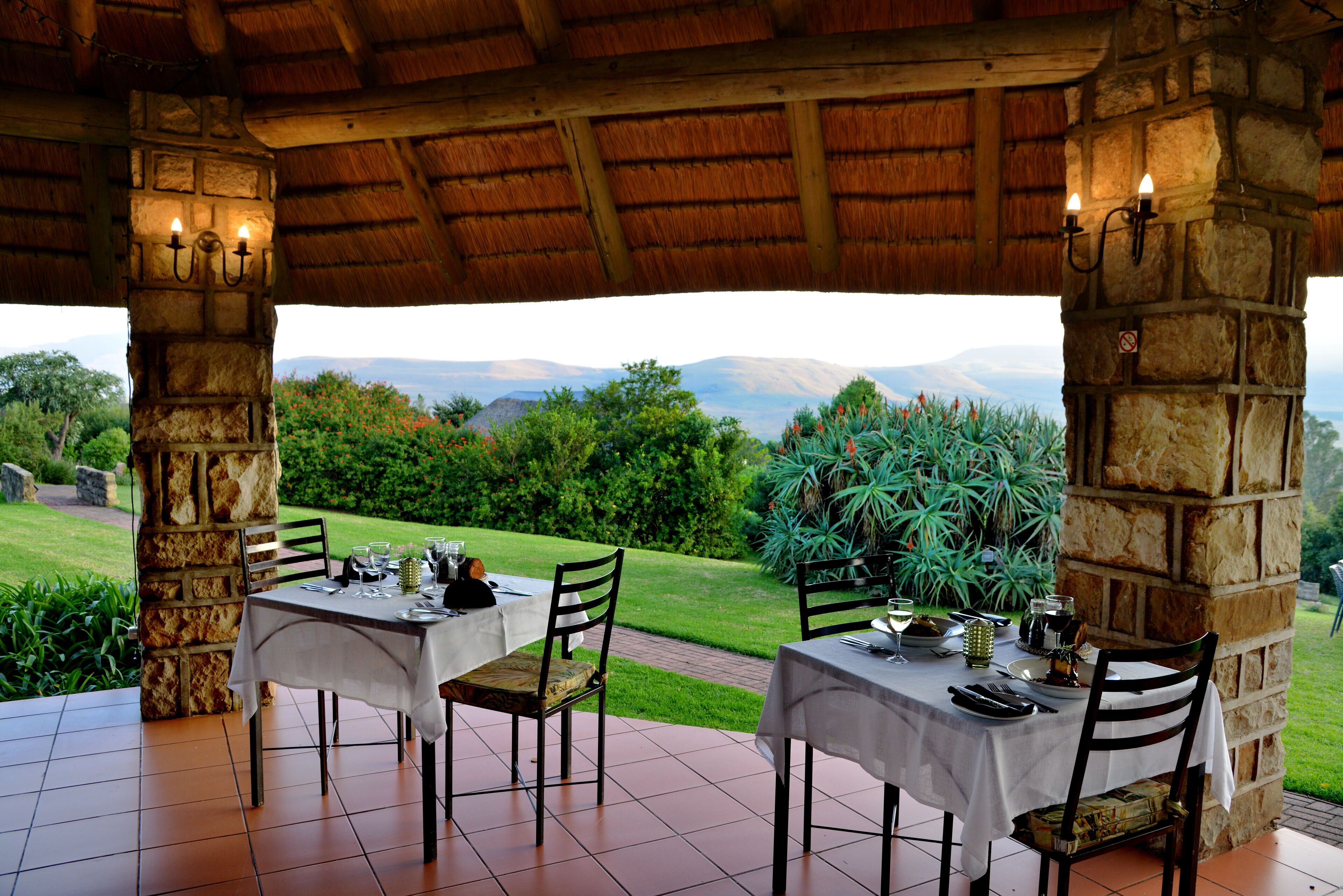A dining area with two tables set for two, each with chairs, under a thatched roof. The tables are covered with white tablecloths and have cutlery and glasses on them. The setting overlooks a lush garden with a view of distant mountains.