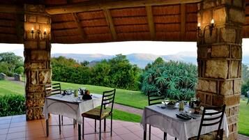 A dining area with two tables set for two, each with chairs, under a thatched roof. The tables are covered with white tablecloths and have cutlery and glasses on them. The setting overlooks a lush garden with a view of distant mountains.