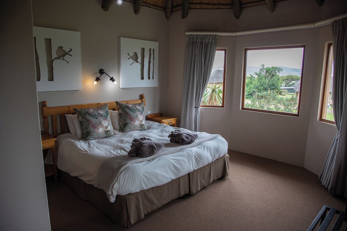 A bedroom with a large bed, two wall-mounted lamps, and a view of greenery through the windows.