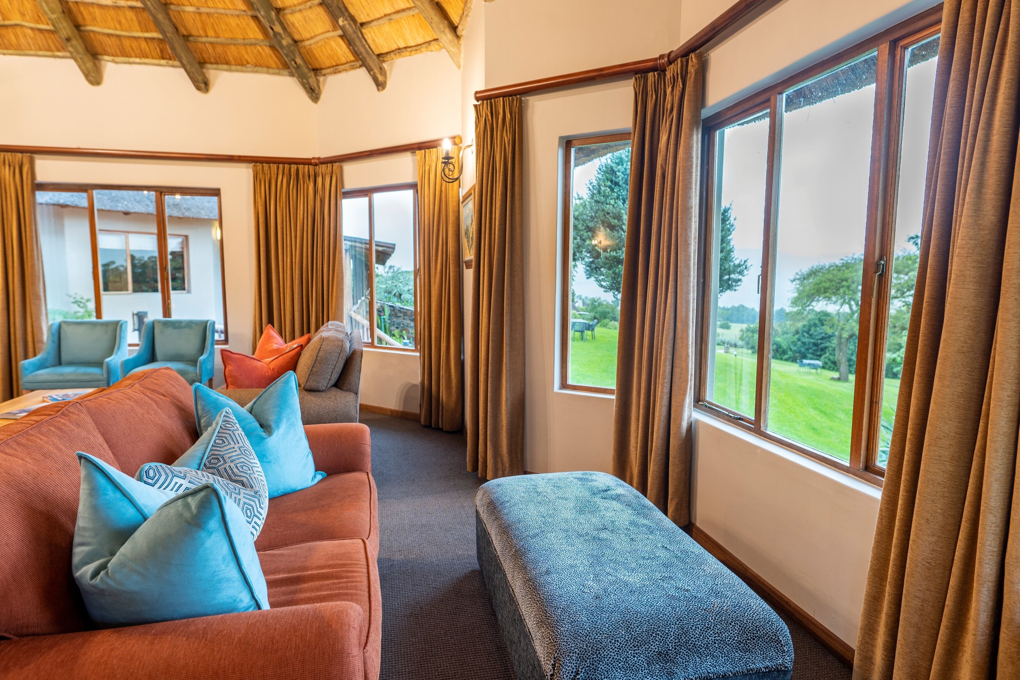 Una habitación acogedora con sofá, reposapiés y sillones, ventanas amplias con cortinas, y vista a zonas verdes.