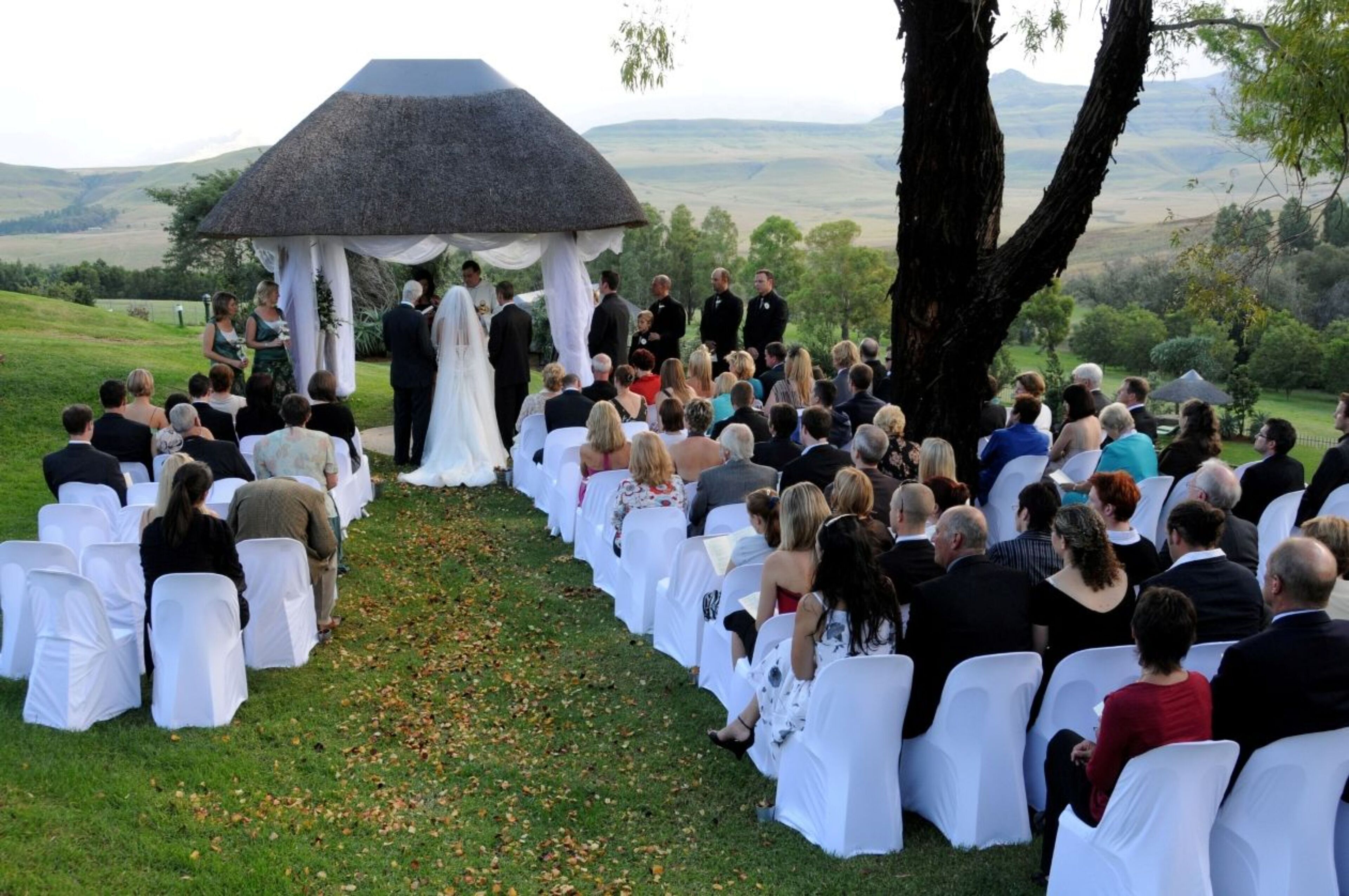 A wedding ceremony outdoors with guests seated on white chairs arranged in rows.