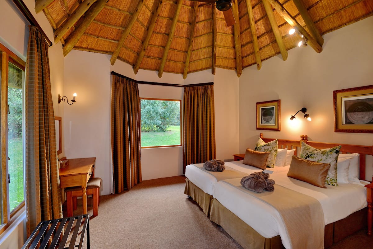 A hotel room with a thatched roof, a bed with multiple pillows, a desk with a chair, and a window with curtains.