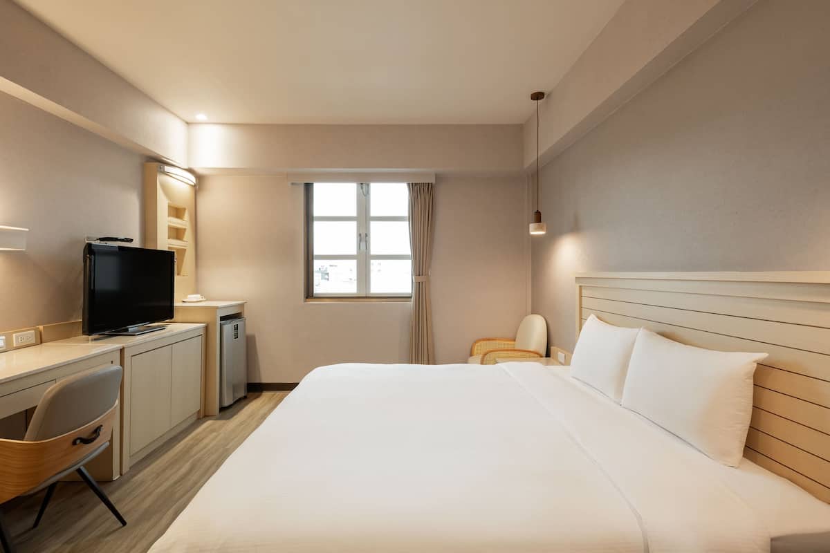 Quarto de hotel com cama, escrivaninha com televisão, cadeira, janela com cortinas e um pequeno frigorífico.