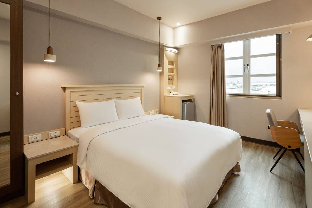 Quarto de hotel bem arrumado, com uma cama grande, mesa de cabeceira e escrivaninha com cadeira.