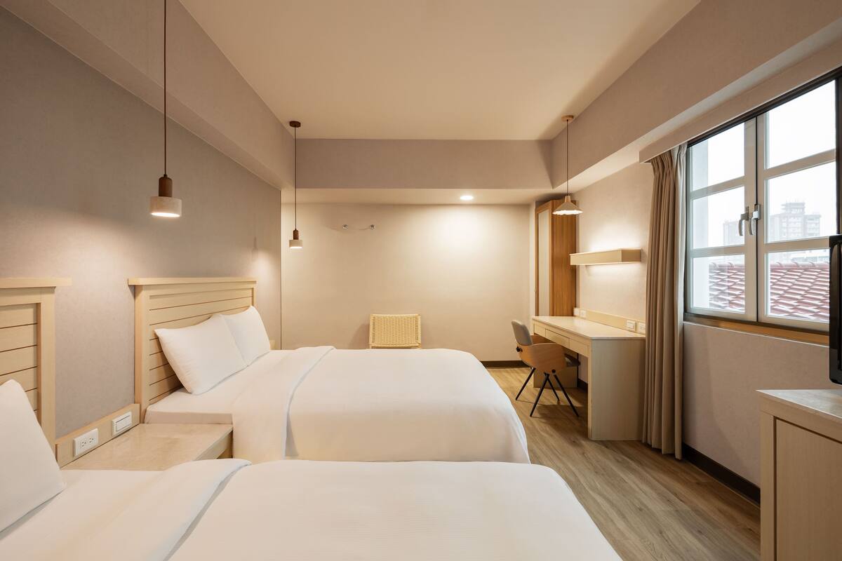 Quarto de hotel com duas camas, uma escrivaninha, uma cadeira, televisão e janela com cortinas.
