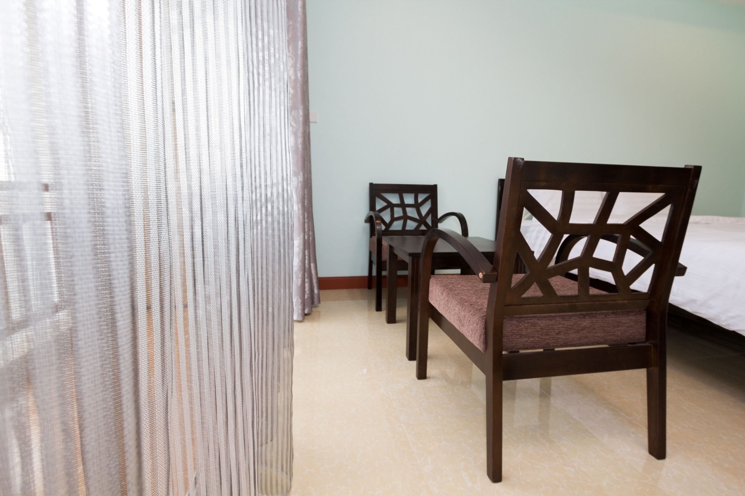 A room with a bed, two wooden chairs, a table, and a sheer curtain.