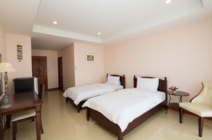 A hotel room with two single beds, a small table with a chair, a desk with a chair, and a lamp.