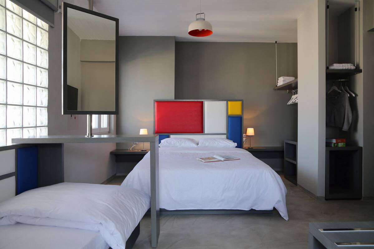 A hotel room with two beds, a large mirror, a red accent wall, and a wardrobe with hanging clothes.