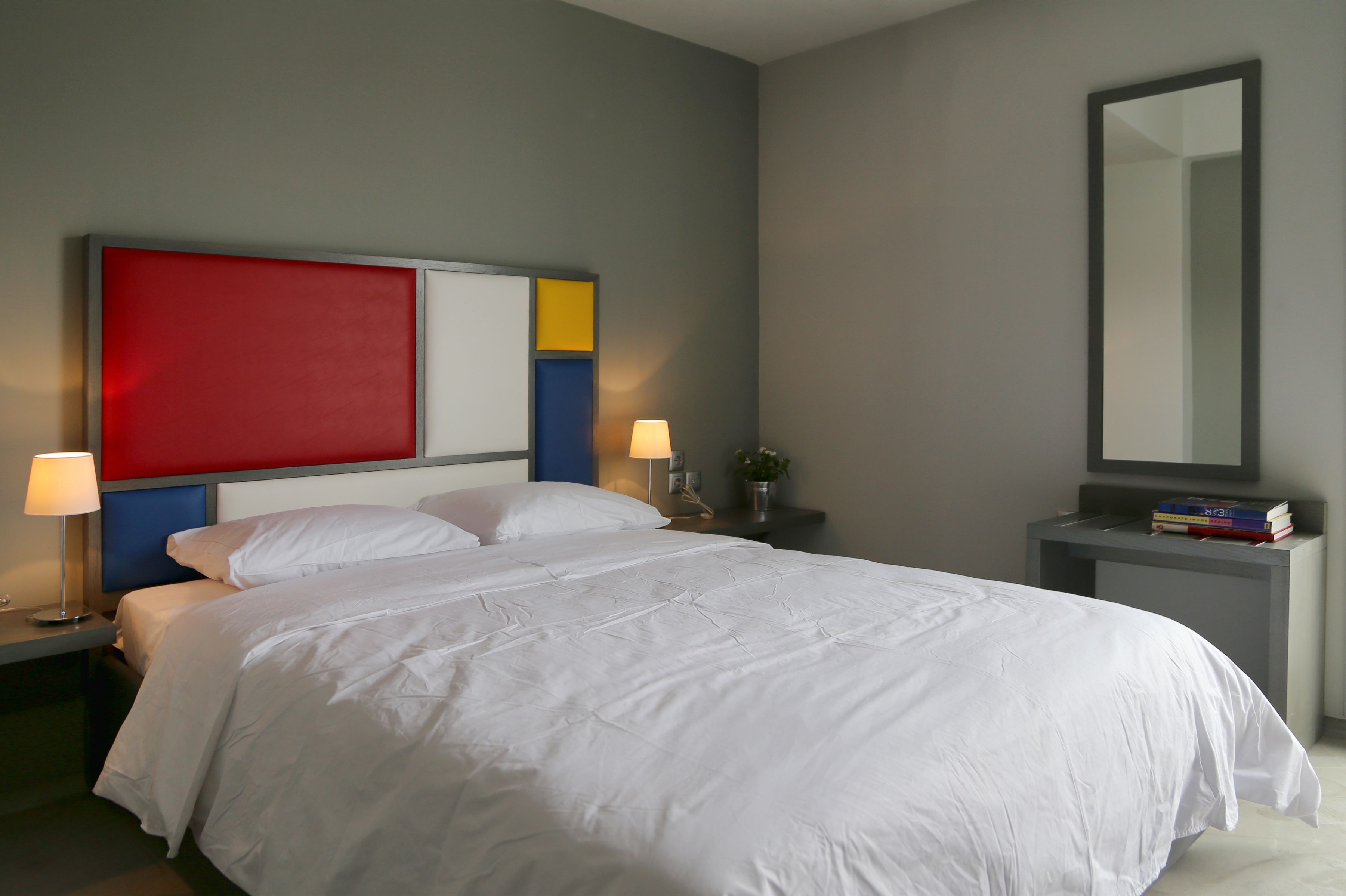Una habitación de hotel moderna con una cama grande, una mesita de noche con lámpara, y un atril con libros y un florero.