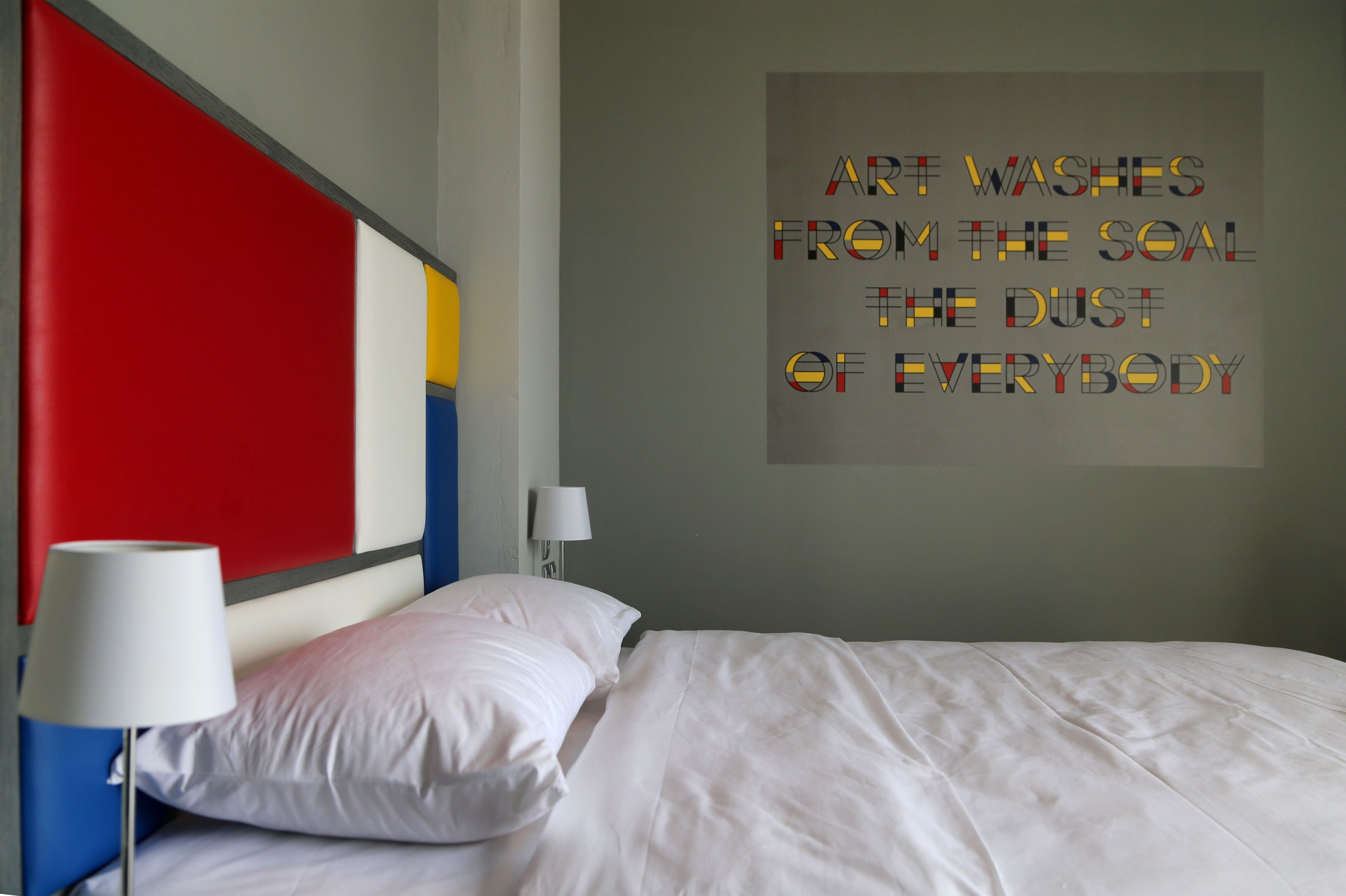 A modern hotel room with a large bed, white bedding, a red accent wall, and a motivational quote on the wall.