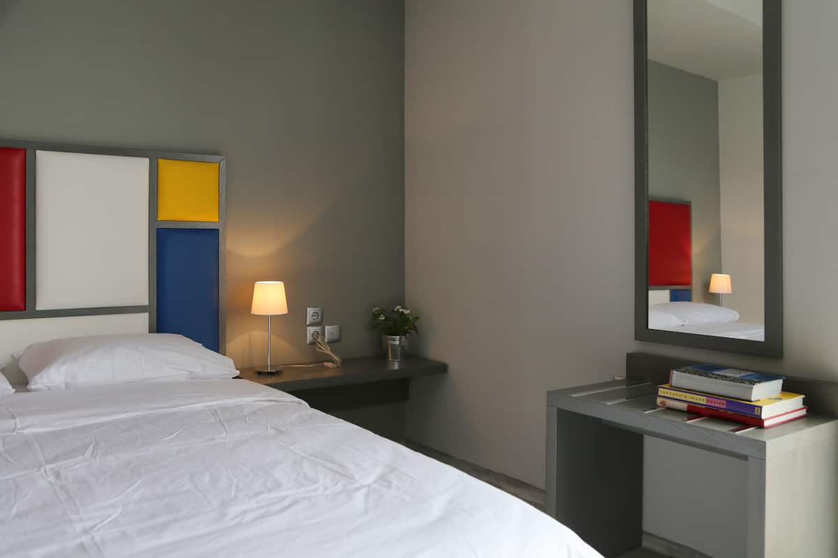 A hotel room with a bed, bedside table, lamp, mirror, and a small desk with books.