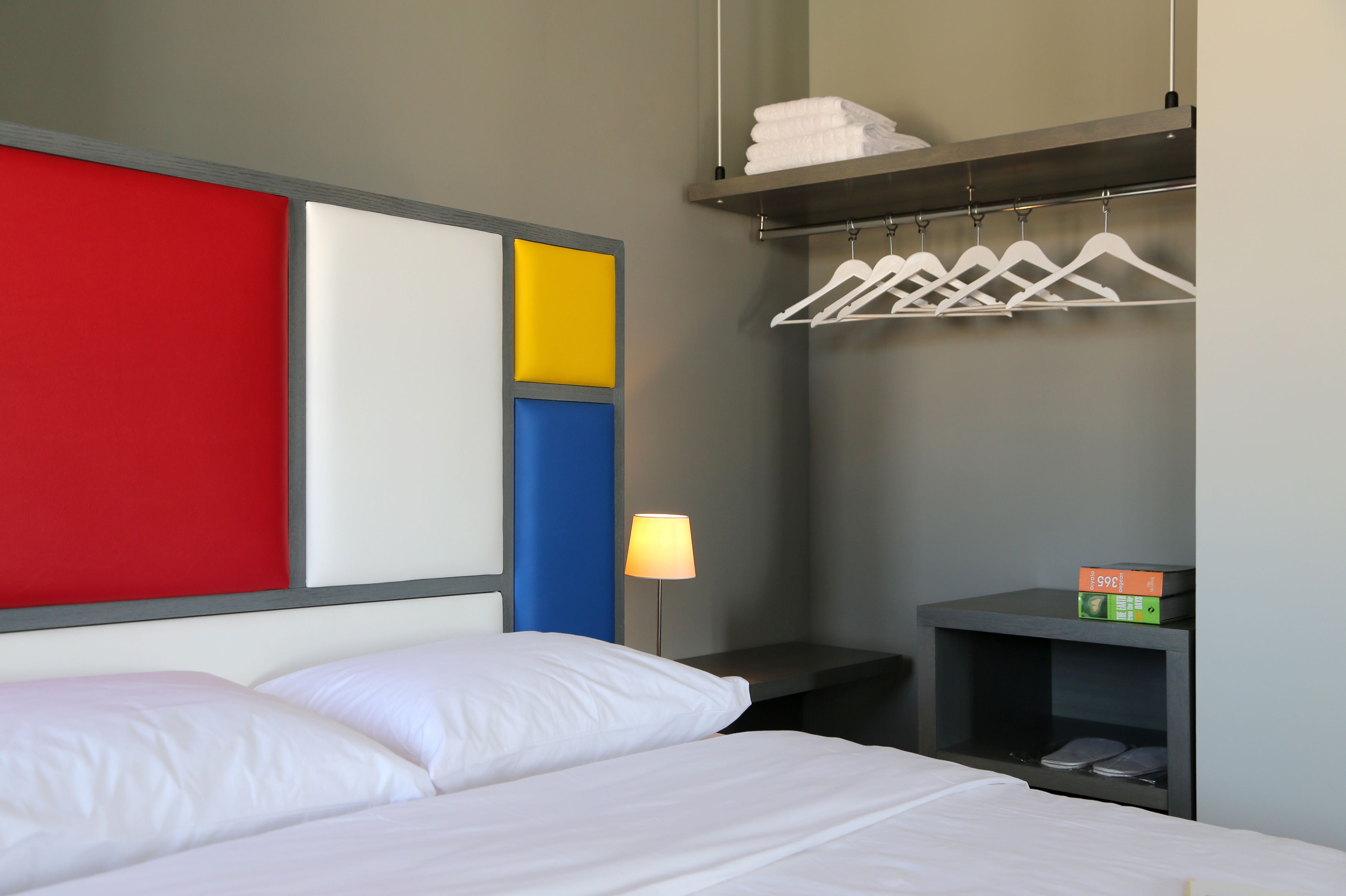 A hotel room with a bed, a nightstand with a lamp, a shelf with towels, and a wall with a red, white, and blue panel.