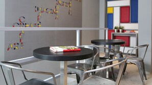 Un café moderno con sillas y mesas de metal, un libro titulado 'Pop Art' sobre una de las mesas, y coloridas obras de arte geométricas en la pared.