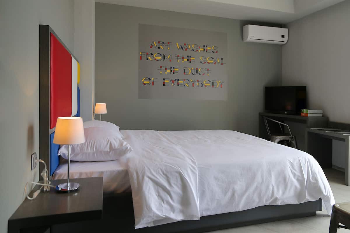 A modern hotel room with a neatly made bed, a bedside table with a lamp, a TV, and a wall-mounted air conditioner.