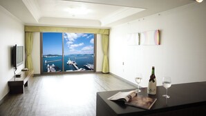 A hotel room with a large window offering a view of the sea and boats. There is a flat-screen TV on a stand, a small table with a bottle of wine and two glasses, and a bench along the wall. - Bay Condo Hotel Tongyeong (Tongyeong)