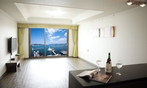 A hotel room with a large window offering a view of the sea and boats. There is a flat-screen TV on a stand, a small table with a bottle of wine and two glasses, and a bench along the wall. - Bay Condo Hotel Tongyeong (Tongyeong)