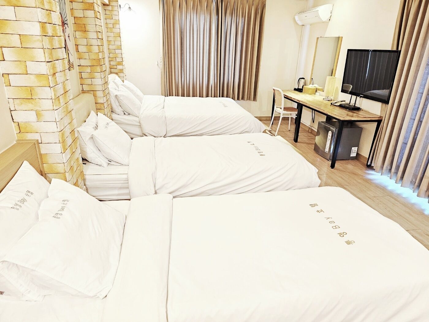 Habitación de hotel con tres camas, un escritorio con silla, un televisor y una pared con diseño de ladrillo.