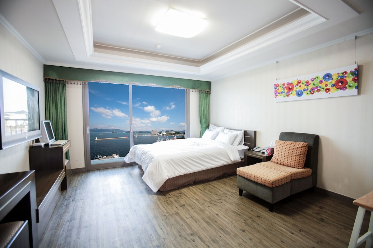 A hotel room with a bed, a desk, a chair, a painting, and a view of the sea.