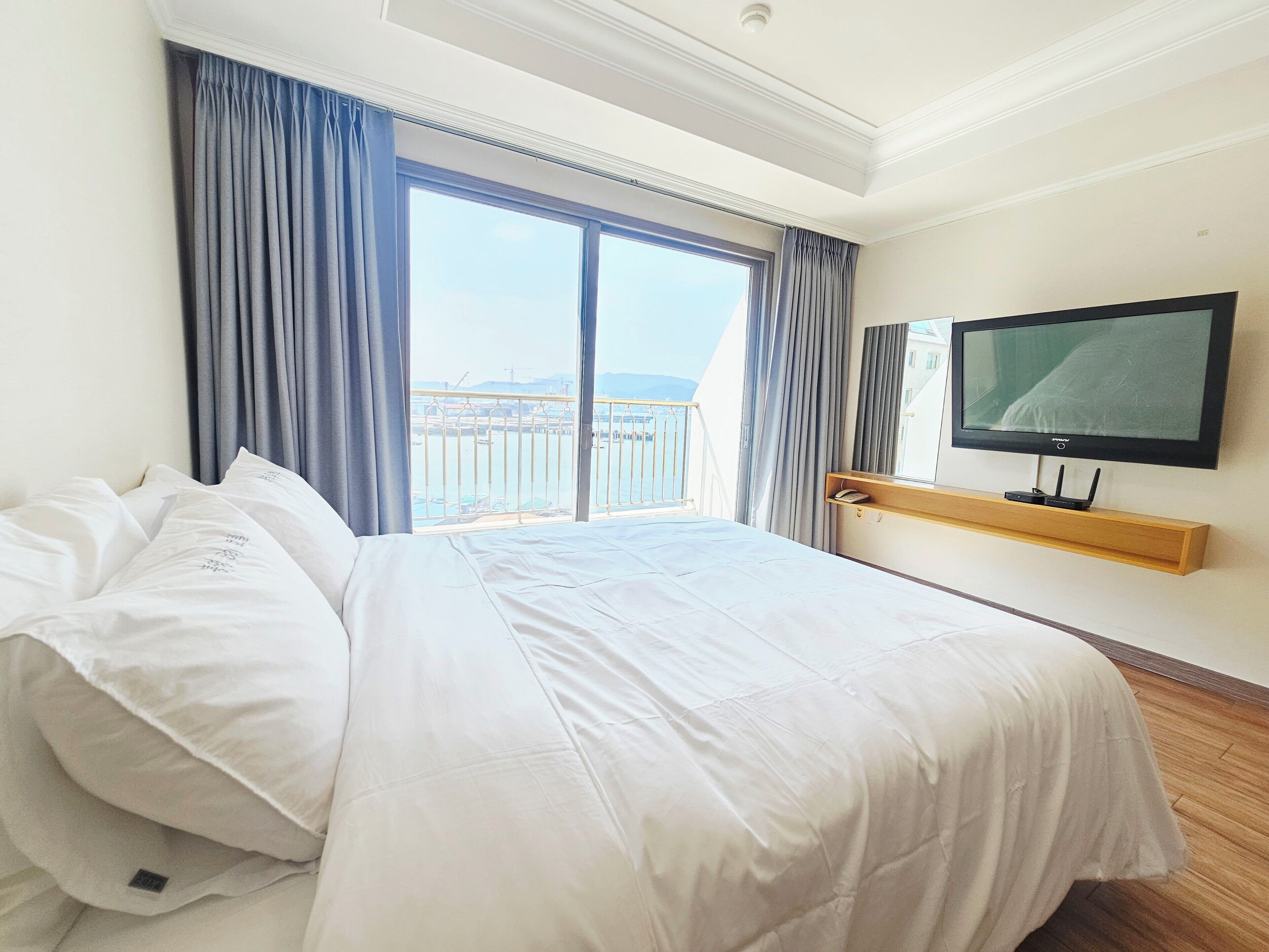 A hotel room with a large bed, a television mounted on the wall, and a balcony with a view of the sea.