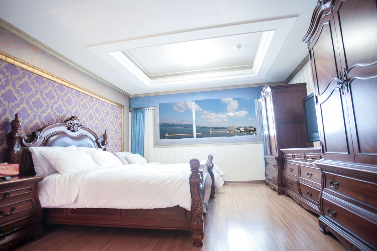 A bedroom with a bed, wooden dresser, and a view of a landscape through a window.