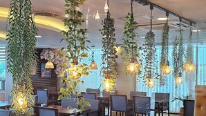 A modern dining area with wooden tables and chairs, hanging plants, and pendant lighting.
