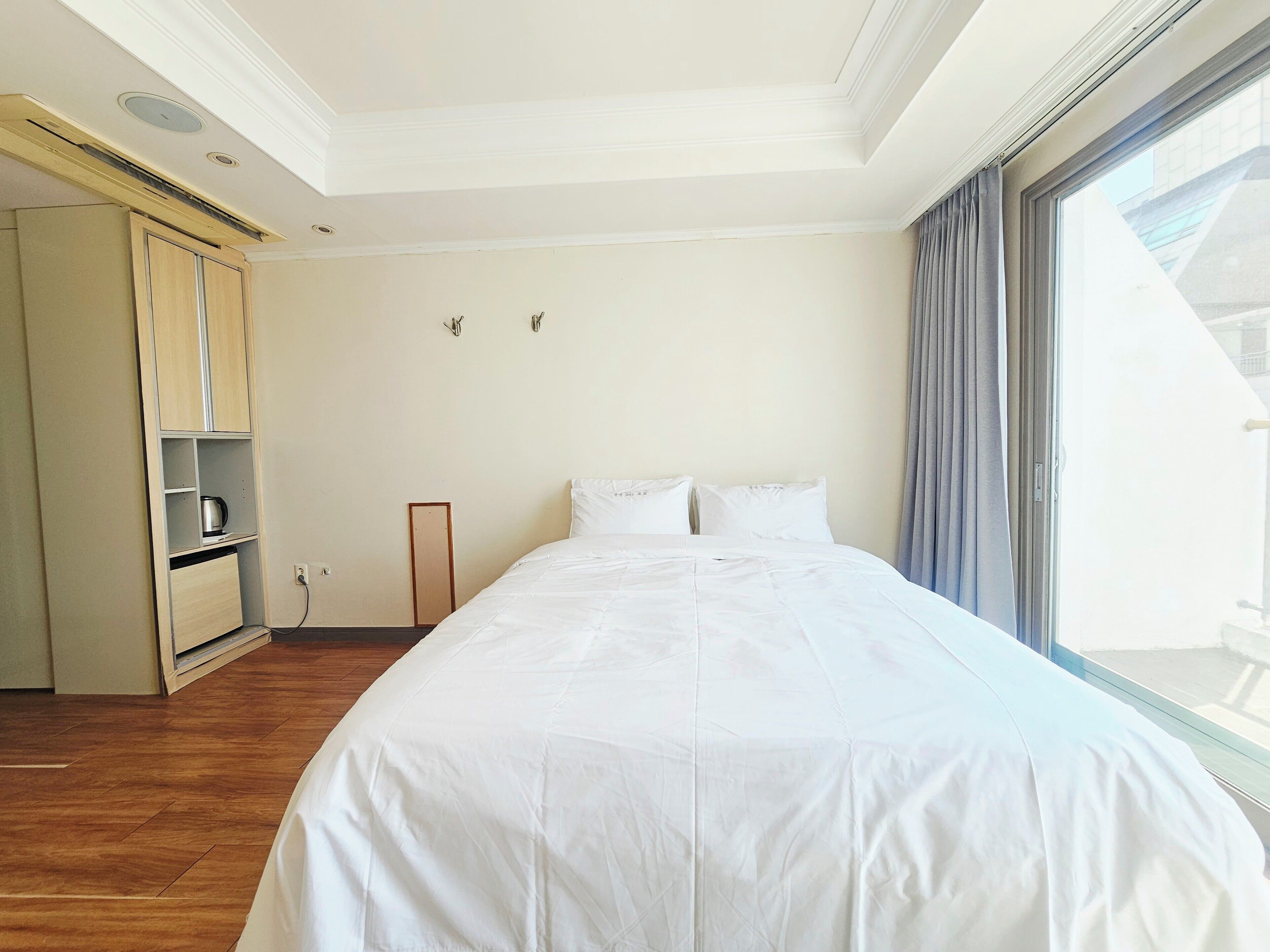 A hotel room with a large bed, a wardrobe, a small shelf with a kettle, and a window with curtains.