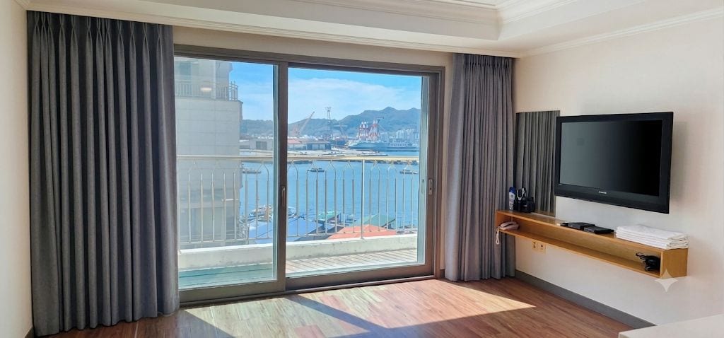 A hotel room with a large sliding glass door leading to a balcony, a flat-screen TV mounted on the wall, and a wooden shelf with a remote control.