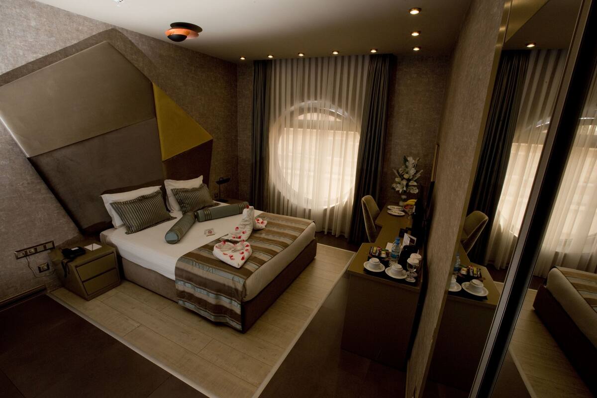 A modern hotel room with a large bed, a desk with a chair, a mirror, and a window with curtains.