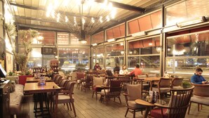 Un restaurante interior con mesas y sillas de madera, ventanas amplias y un techo con luces colgantes.