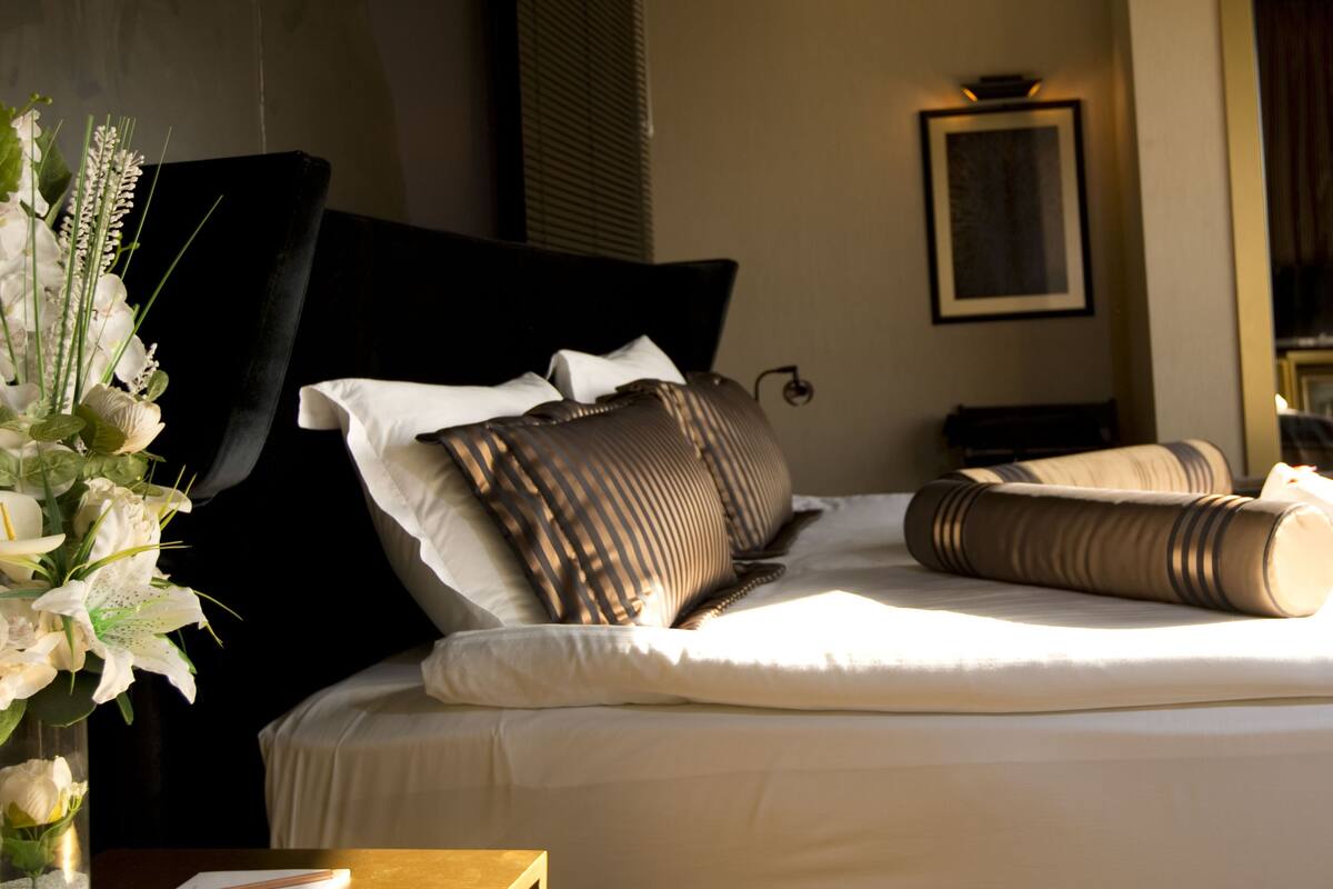 A neatly made bed with white linens and multiple pillows, a vase with white flowers, and a framed picture on the wall.