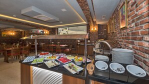 Zona de buffet con diferentes opciones de comida, pared de ladrillo y comedor con mesas y sillas.